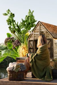 A woman wrapped in a blanket, surrounded by seasonal produce and greenery.